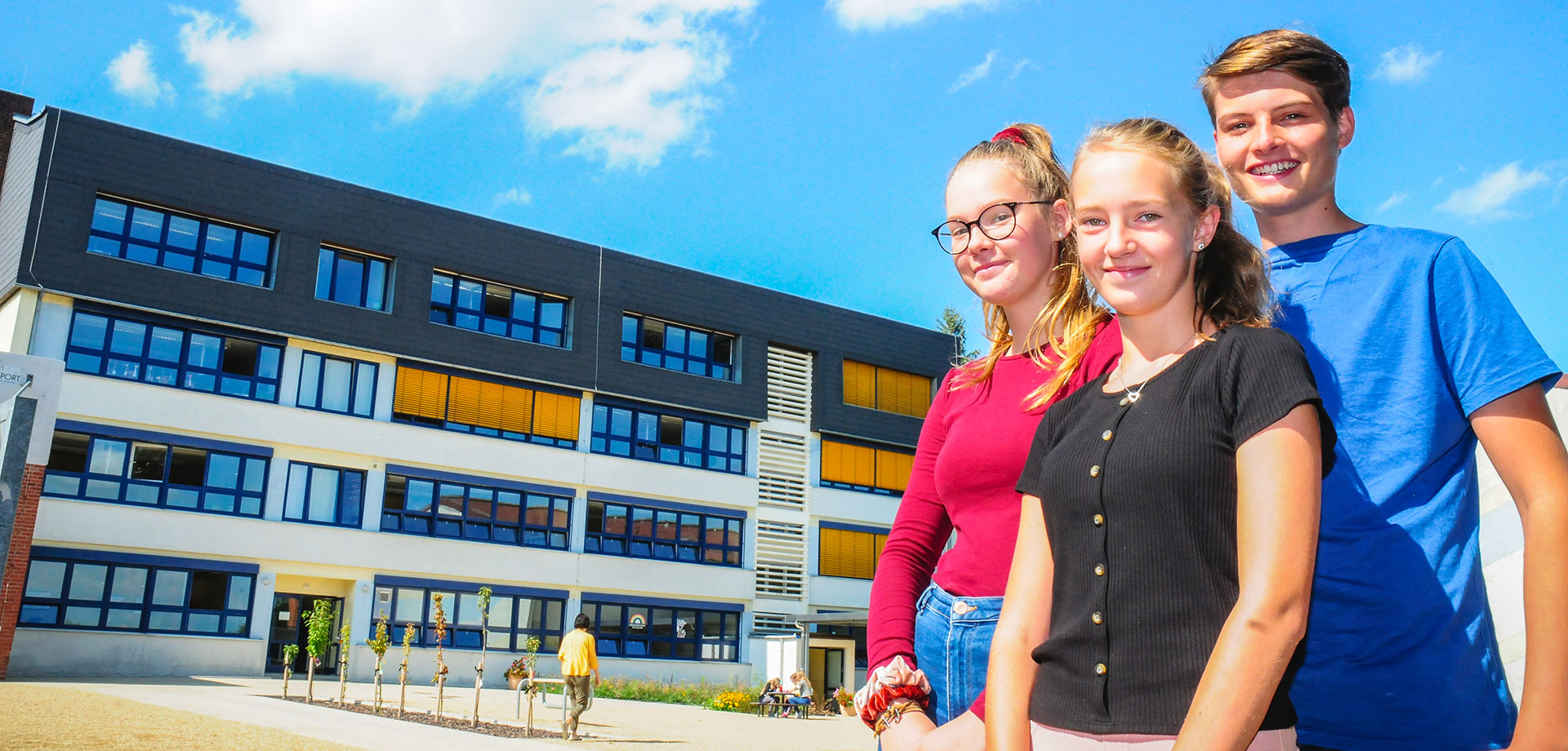 DPFA-Regenbogen-Gymnasium Augustusburg: Zwei Mädchen und ein Junge stehen auf dem Schulhof mit dem Schulgebäude im Hintergrund.
