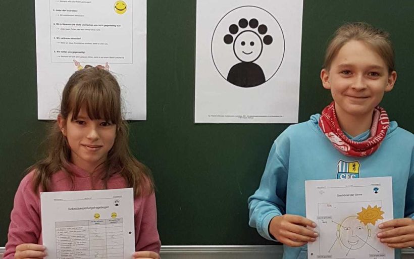 Ein Mädchen und ein Junge stehen in ihrem Klassenzimmer vor einer Tafel und halten zwei Arbeitsblätter in den Händen.
