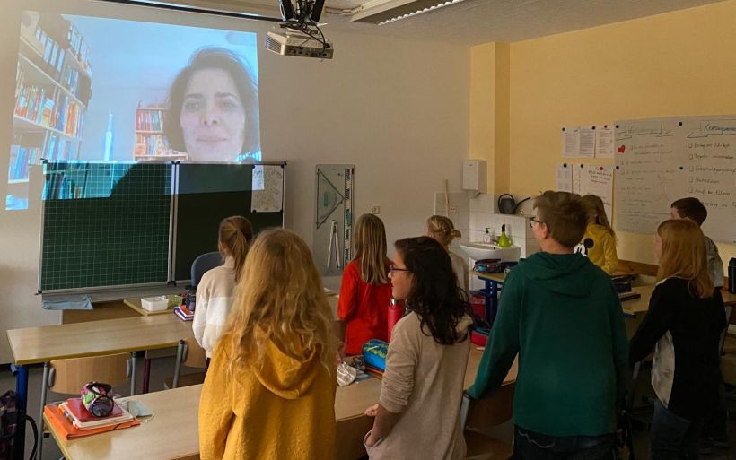 Schüler stehen in einme Klassenzimmer und blicken auf ihre Lehrerin, die über eine Linwand digital zugeschaltet ist.