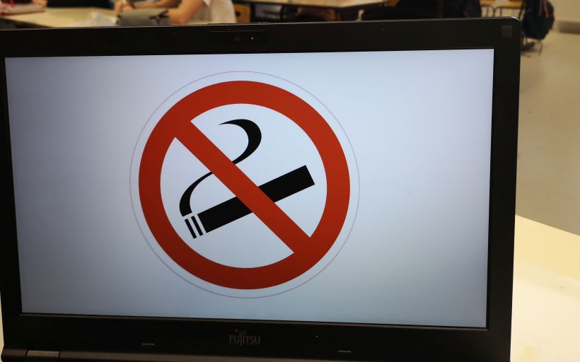 Auf einem Laptop ist ein Rauchverbotsschild zu sehen, dahinter Schüler, die in einem Klassenzimmer sitzen.