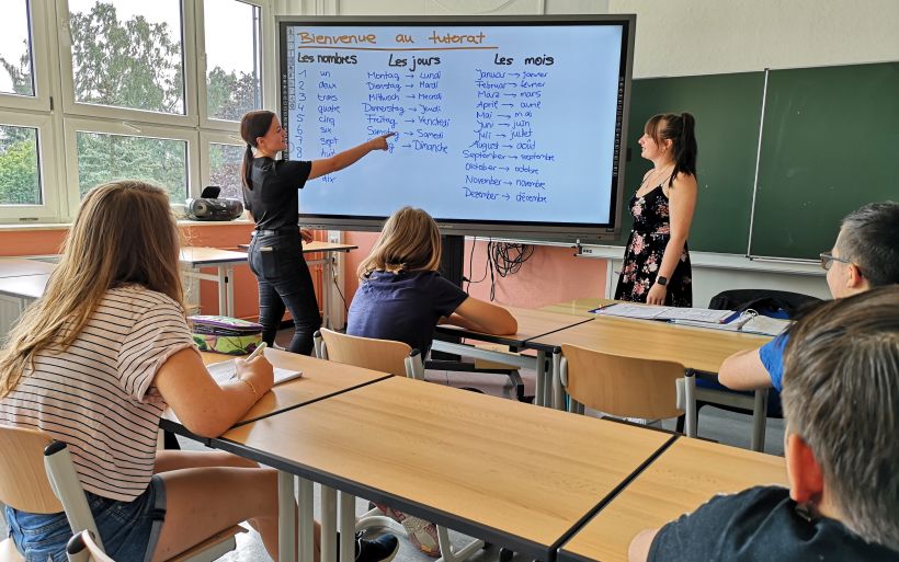 Schüler sitzen in einem Klassenzimmer und schauen auf eine digitale Tafel, an der eine Schülerin etwas erklärt.