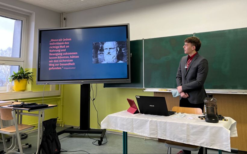 Ein junger Mann hält einen Vortrag vor einer Klasse. Er nutzt die digitale Tafel als Hilfsmittel.