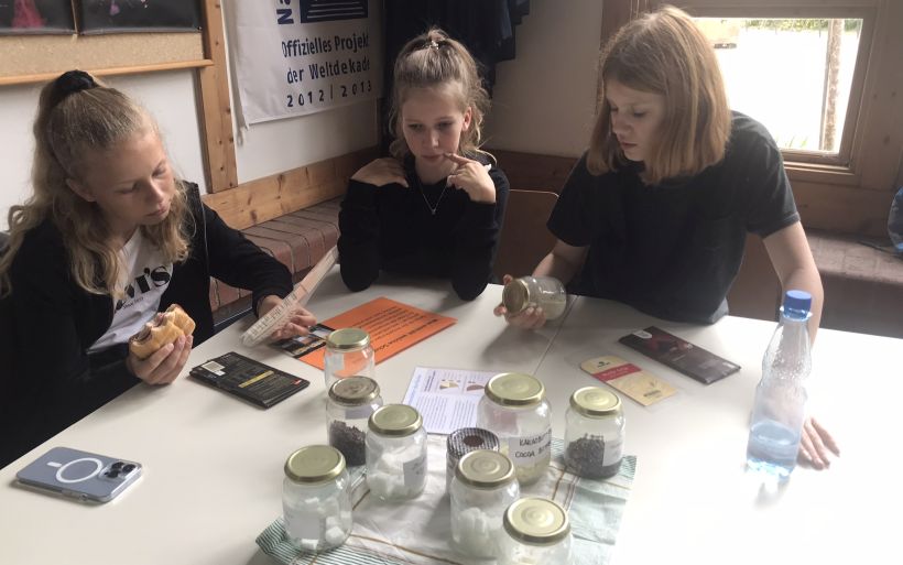 Drei Mädchen sitzen an einem Tisch und schauen sich Dinge zum Thema Kakao-Herstellung an.