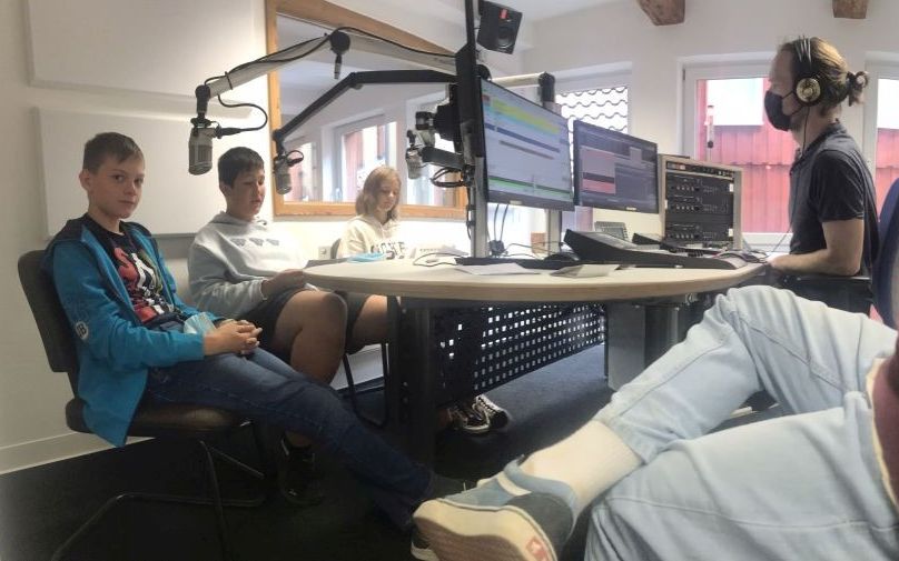 Drei Schüler sitzen mit einem Radiomoderator in einem Radio-Studio und nehmen einen Beitrag auf.