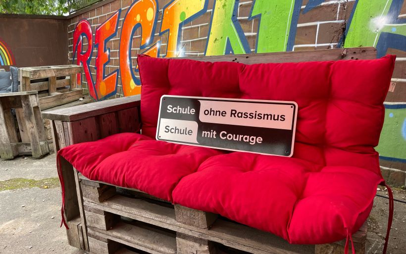 Das offizielle Schild vom Netzwerk "Schule ohne Rassismus" steht dekorativ auf einem roten Sofa.