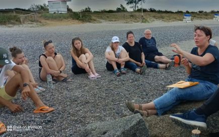 eine Gruppe Jugendliche sitzt am Strand im Kreis. Eine Lehrerin erklärt etwas.