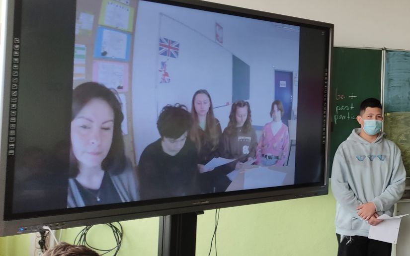 Schüler steht neben einer digitalen Tafel, auf der eine Videokonferenz mit anderen Schülern läuft. Er hält einen Vortrag.